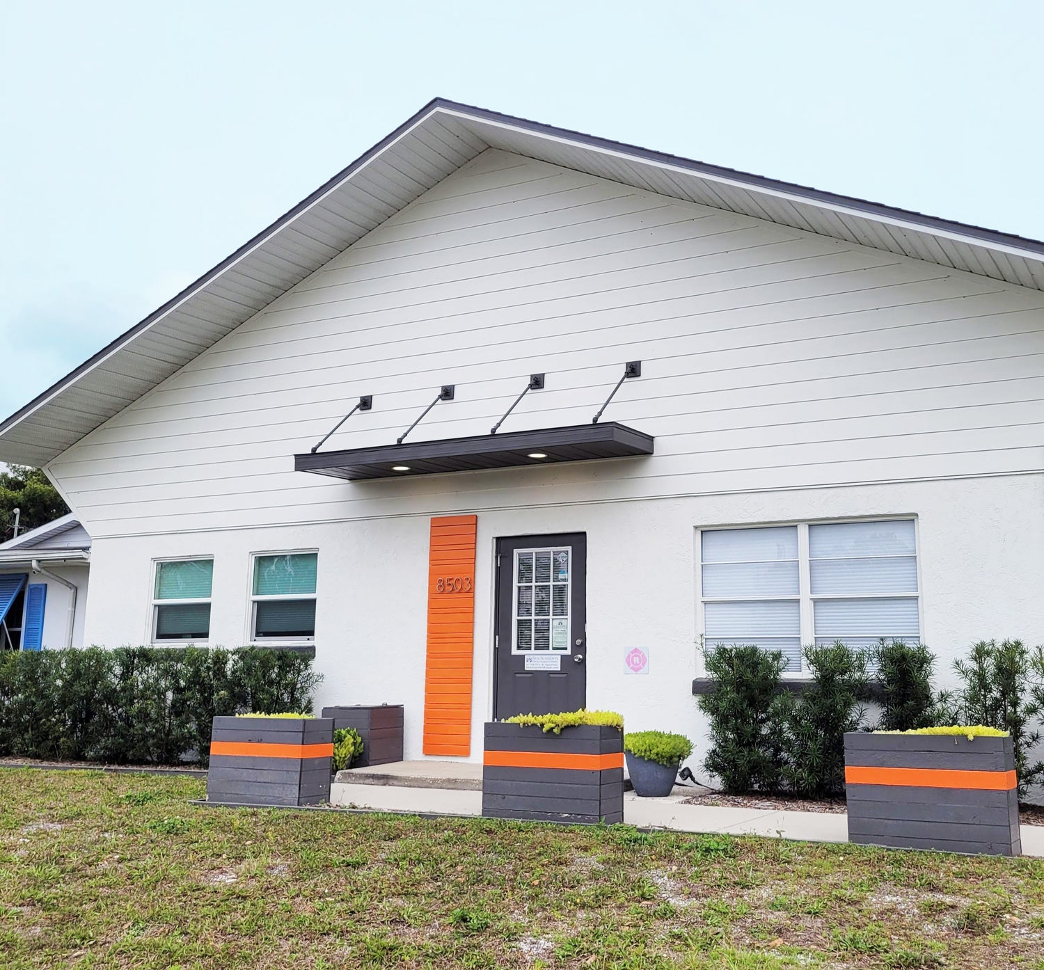 Picture of the front of the Beachcombers henna studio building. It's a white building with a gray awning and door and orange accents..