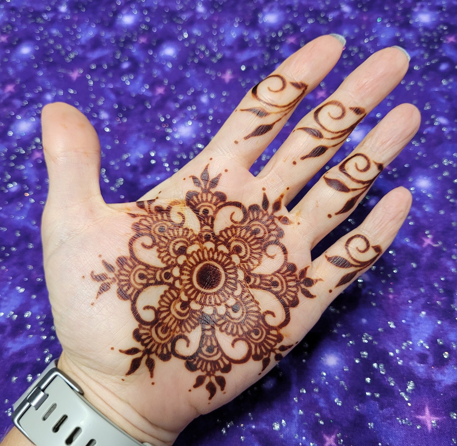 A dark henna stain of a mandala on the palm with a purple background.