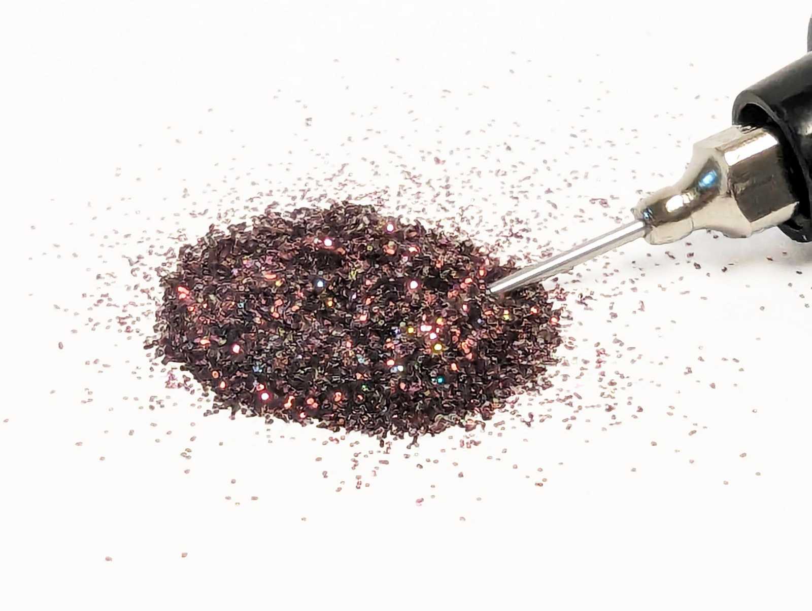 Metallic burgundy glitter being dispensed from a small handheld tool on a white background