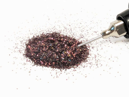 Metallic burgundy glitter being dispensed from a small handheld tool on a white background