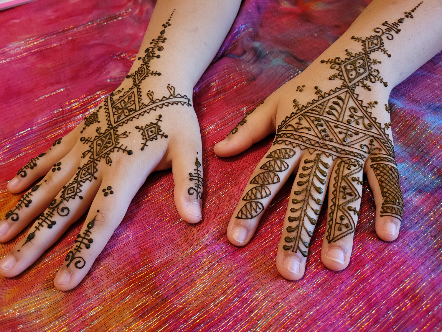 Two tops of hands with Fessi Moroccan style henna designs on a bright pink background