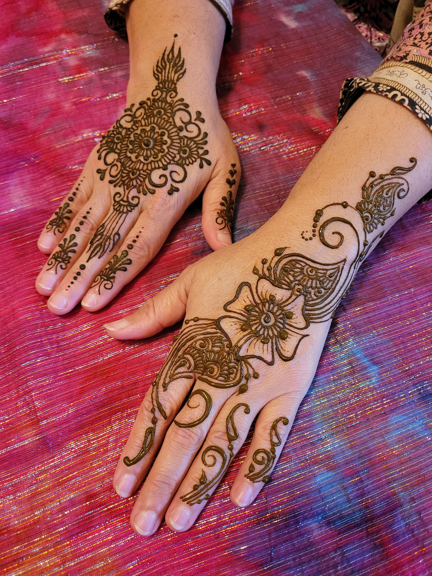 Two hands with henna on a pink background. One hand has a mandala and the other has a sangeet strips tyle. both are intricate designs