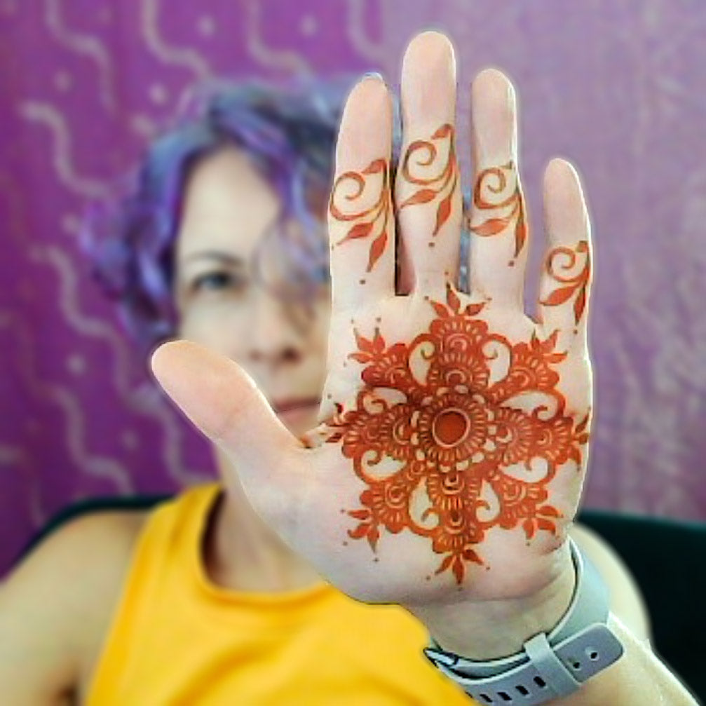 Picture of Jody holding up her palm to the camera slightly in front of her face. The palm has a fresh orange henna stain.